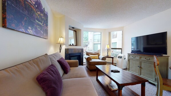 A living room with a sofa, armchair, coffee table, TV, fireplace, and large windows. - Marquise on Blackcomb, Pool & Hot Tub, Easy Ski Access, Free WiFi (Whistler)