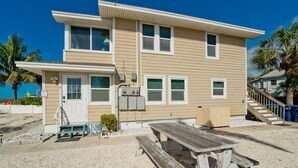 A two-story building with a front porch, a small patio with a wooden table and benches, and a view of the beach.
