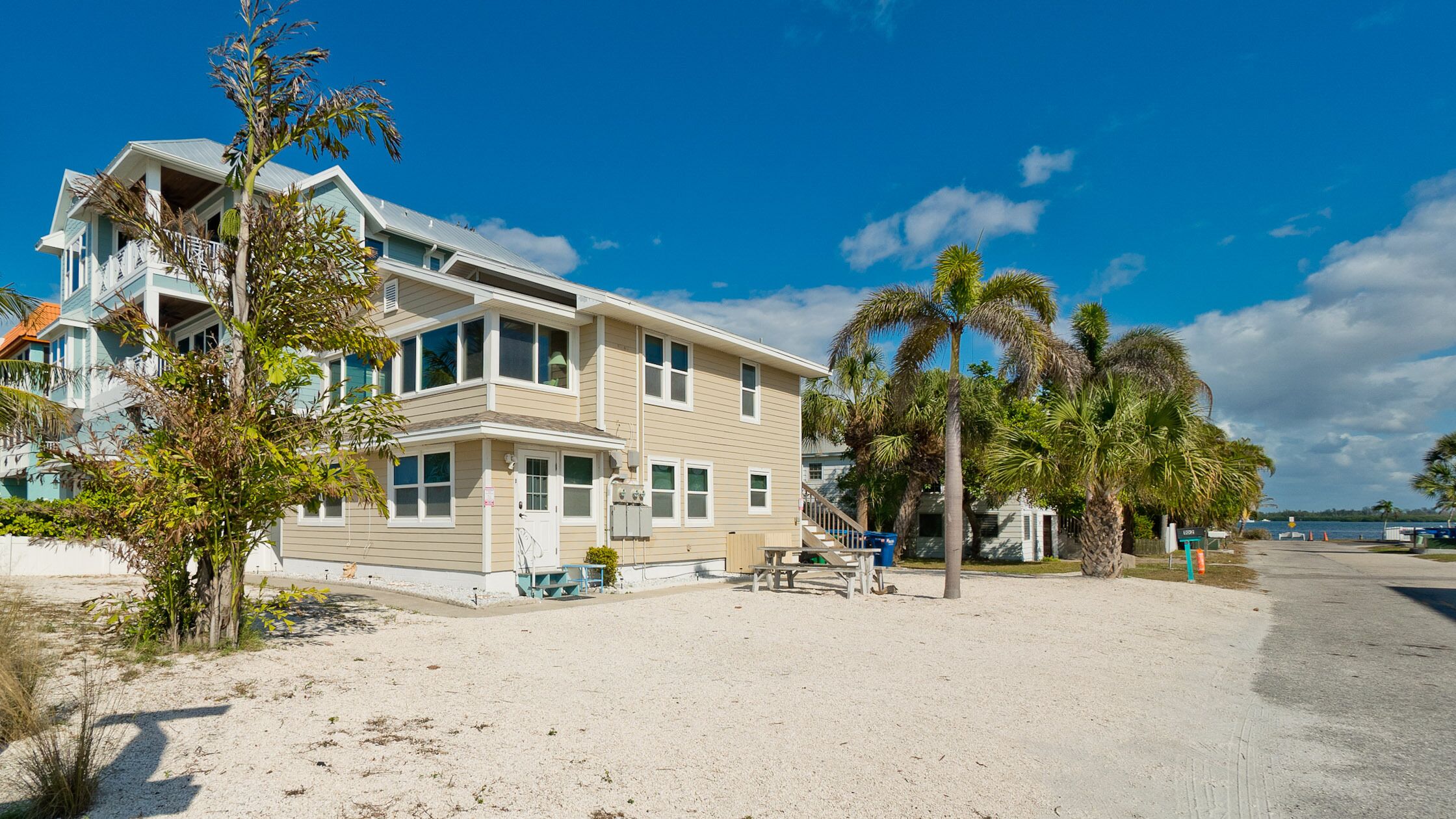 A two-story house with a balcony, located on a sandy beach with palm trees.