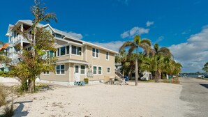 A two-story house with a balcony, located on a sandy beach with palm trees.