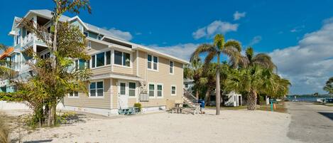 A two-story house with a balcony, located on a sandy beach with palm trees.