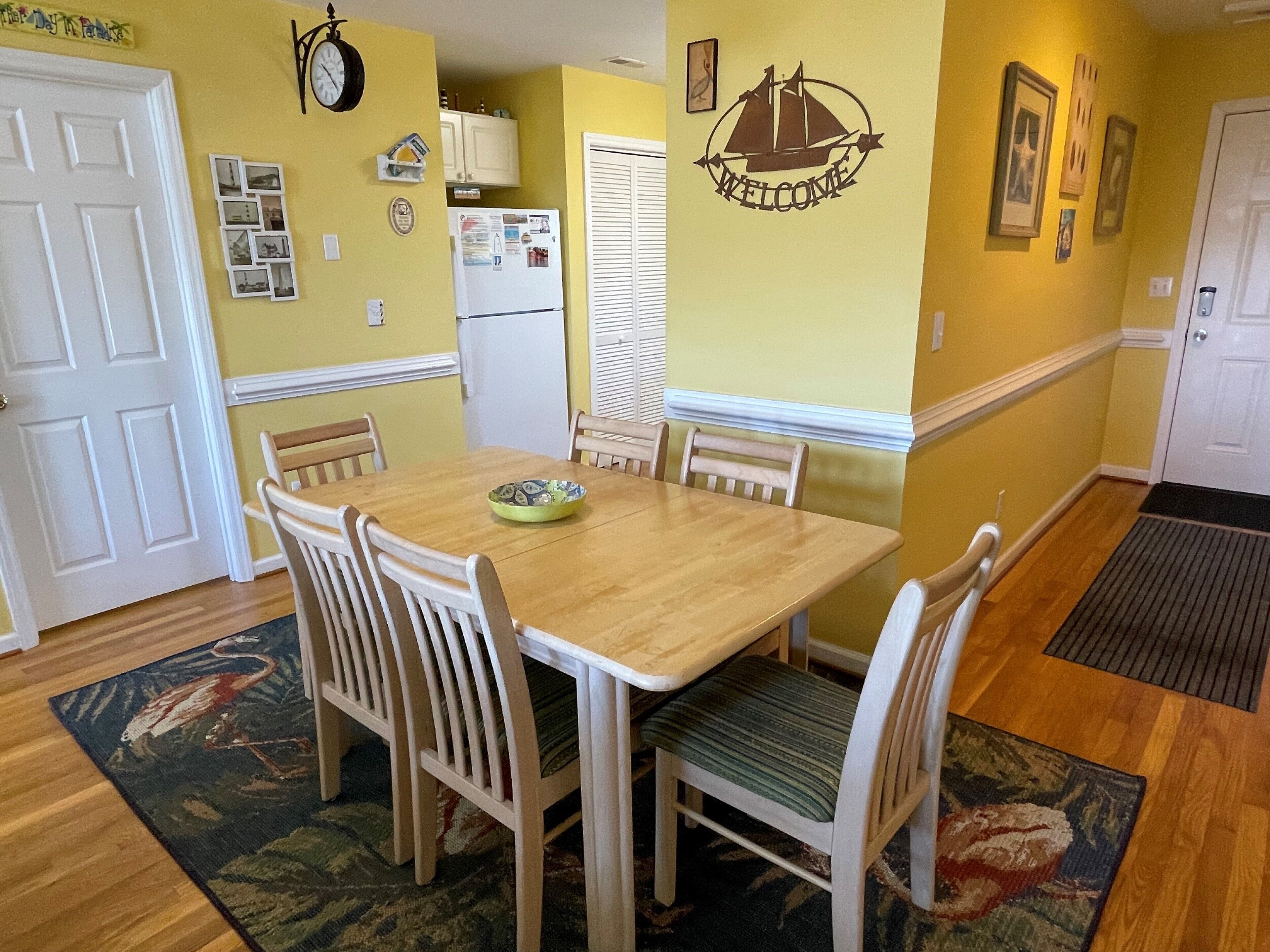 A dining area with a wooden table and chairs, a clock on the wall, and a welcome sign.