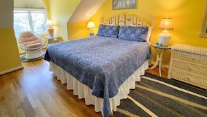 Ein Schlafzimmer mit einem großen Bett, zwei Nachttischlampen, einem Sessel, einem kleinen Tisch und einem Fenster mit Blick auf Bäume.