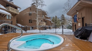A wooden deck with a hot tub, surrounded by snow-covered buildings and a metal fence.
