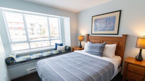 Een slaapkamer met een groot raam, een bed met blauw beddengoed, twee bedlampjes en een ingelijst schilderij aan de muur.