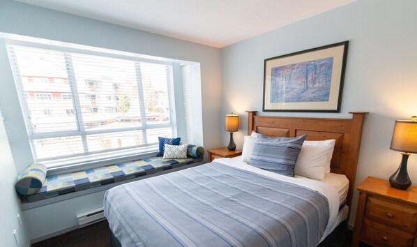 Un dormitorio con un ventanal grande, una cama con ropa de cama azul, dos lámparas de noche y un cuadro enmarcado en la pared.