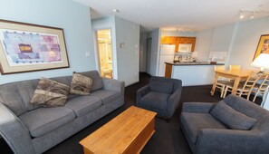 A living room with a grey sofa, two armchairs, a wooden coffee table, and a dining area with a wooden table and chairs.