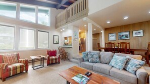 A spacious living area with a high ceiling, large windows, a dining area, and a kitchen.