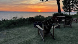 A sunset view with a wooden table and chairs, a fire pit, and a grassy area by the water. - Lakefront Home in the Heart of Aurora, Sunset Ready! (Aurora)