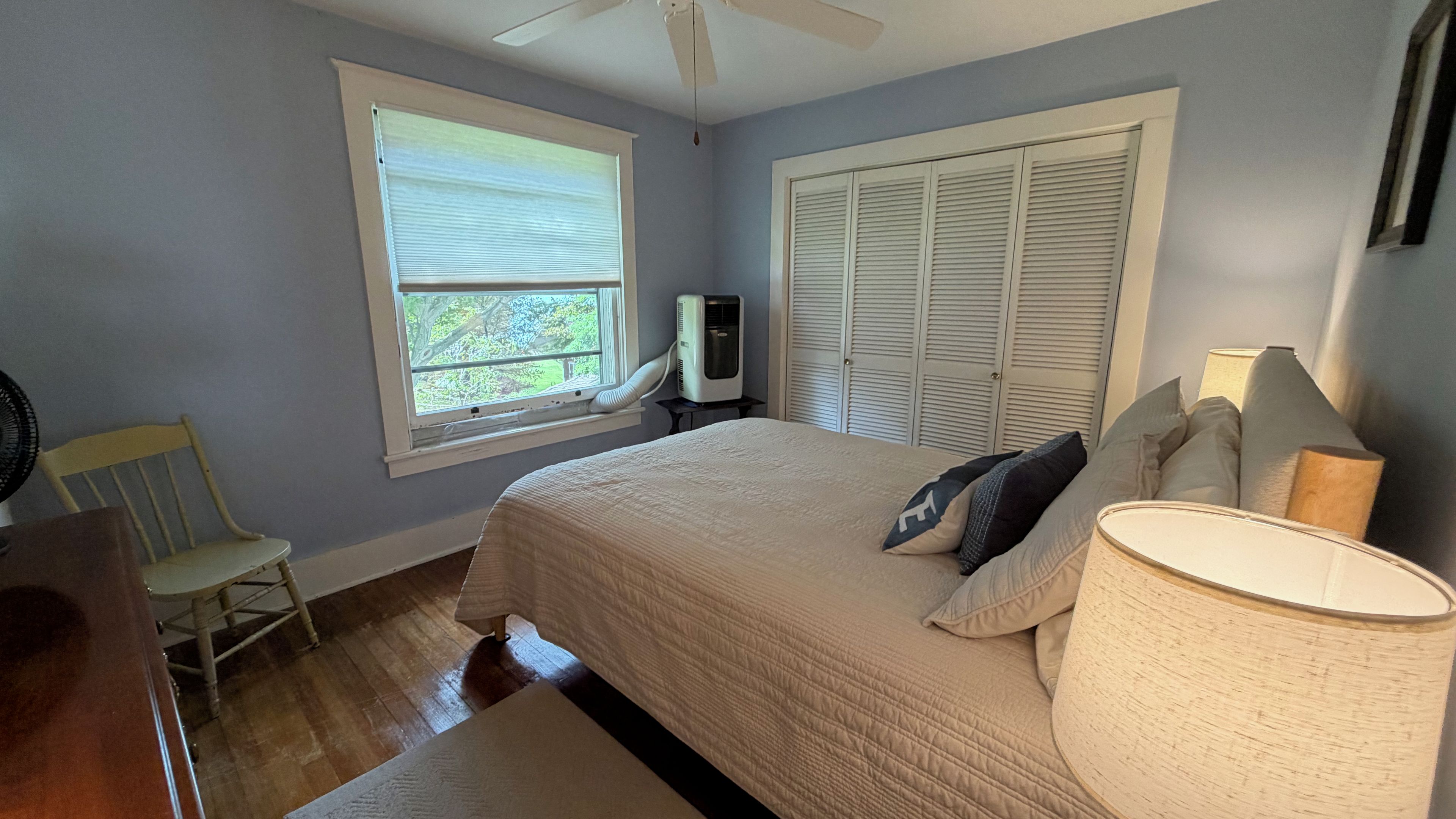 A bedroom with a bed, a window, a heater, a chair, and a lamp.