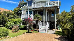 A two-story house with a balcony, a covered porch, and a garden with various plants and flowers. - Island Rest:  Marsh and distant sound views on Jackson Circle (Ocracoke)