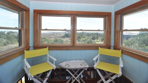 A room with a view through large windows, a small table, and two yellow chairs. - Island Rest:  Marsh and distant sound views on Jackson Circle (Ocracoke)