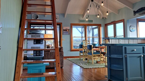 A spacious room with a wooden staircase leading to an upper level, large windows, and a kitchen area with blue cabinets. - Island Rest:  Marsh and distant sound views on Jackson Circle (Ocracoke)