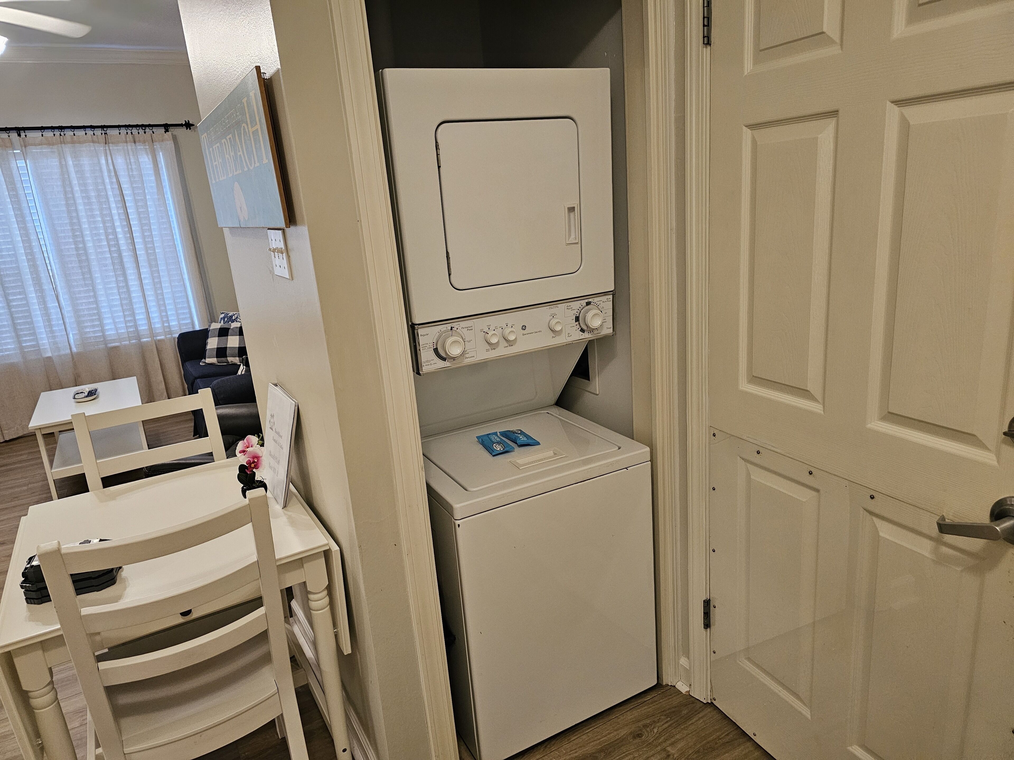A compact laundry room with a washing machine and dryer, a small table with a chair, and a door leading to another room.