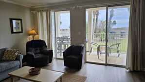 Ein Wohnzimmer mit einer Couch, zwei Sesseln, einem Couchtisch und Blick auf einen umzäunten Außenbereich mit Stühlen und einem Tisch.
