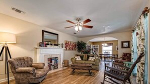 A living room with a fireplace, a ceiling fan, and a dining area with chairs and a table. - A perfect home for your vacation! (Waco)