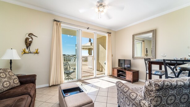 A living room with a sofa, ottoman, and a TV stand. There is a balcony with a view of the outside. - The Inn at Dauphin Island 106 1BR Walks right out to Pools and Beach! (Dauphin Island)