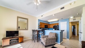 A living room with a TV, sofa, and ottoman, a dining area with chairs and a table, and a kitchen with wooden cabinets and a refrigerator. - The Inn at Dauphin Island 106 1BR Walks right out to Pools and Beach! (Dauphin Island)