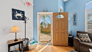 Una habitación con puerta de madera, perchero, mesa con lámpara, sofá y vista a un jardín a través de la puerta.