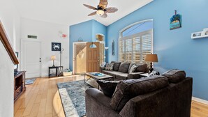 Una sala de estar con pared azul, ventilador de techo, sofá, mesa de centro y puerta que da a otra habitación.