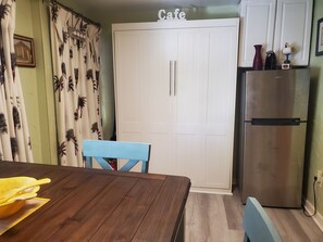 A compact kitchen with a dining table, a blue chair, a white door with a 'Cafe' sign, and a stainless steel refrigerator. - STEPS FROM BEACH, DAILY RENTALS,PET AND FAMILY FRIENDLY BEACH HOUSE (Clearwater Beach)