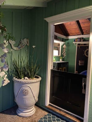 Uma porta verde com um detalhe decorativo branco, um vaso com planta e uma pequena estante com livros e objetos decorativos.