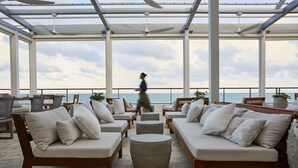 A covered outdoor seating area with sofas and round tables, overlooking the sea.