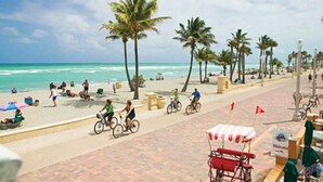 A beach scene with palm trees, a paved walkway, and people relaxing on the sand.