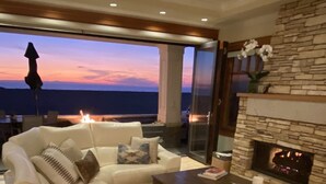 A modern living room with a white leather sofa, a fireplace, and a view of the ocean at sunset. - Luxury Oceanfront Home with Rooftop Deck, Hot Tub, Fire Pit (Newport Beach)