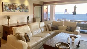 A spacious living room with a large beige sofa, a wooden coffee table, and a view of the sea through a large window. - Luxury Oceanfront Home with Rooftop Deck, Hot Tub, Fire Pit (Newport Beach)