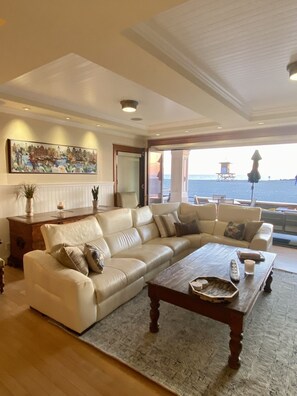 A spacious living room with a large beige sofa, a wooden coffee table, and a view of the sea through a large window. - Luxury Oceanfront Home with Rooftop Deck, Hot Tub, Fire Pit (Newport Beach)