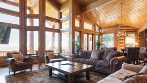 A spacious log cabin interior with large windows, a wooden ceiling, and a cozy seating area with a sofa and armchairs. - Deer Valley Cabin W/ Hot Tub | Theater (Park City)