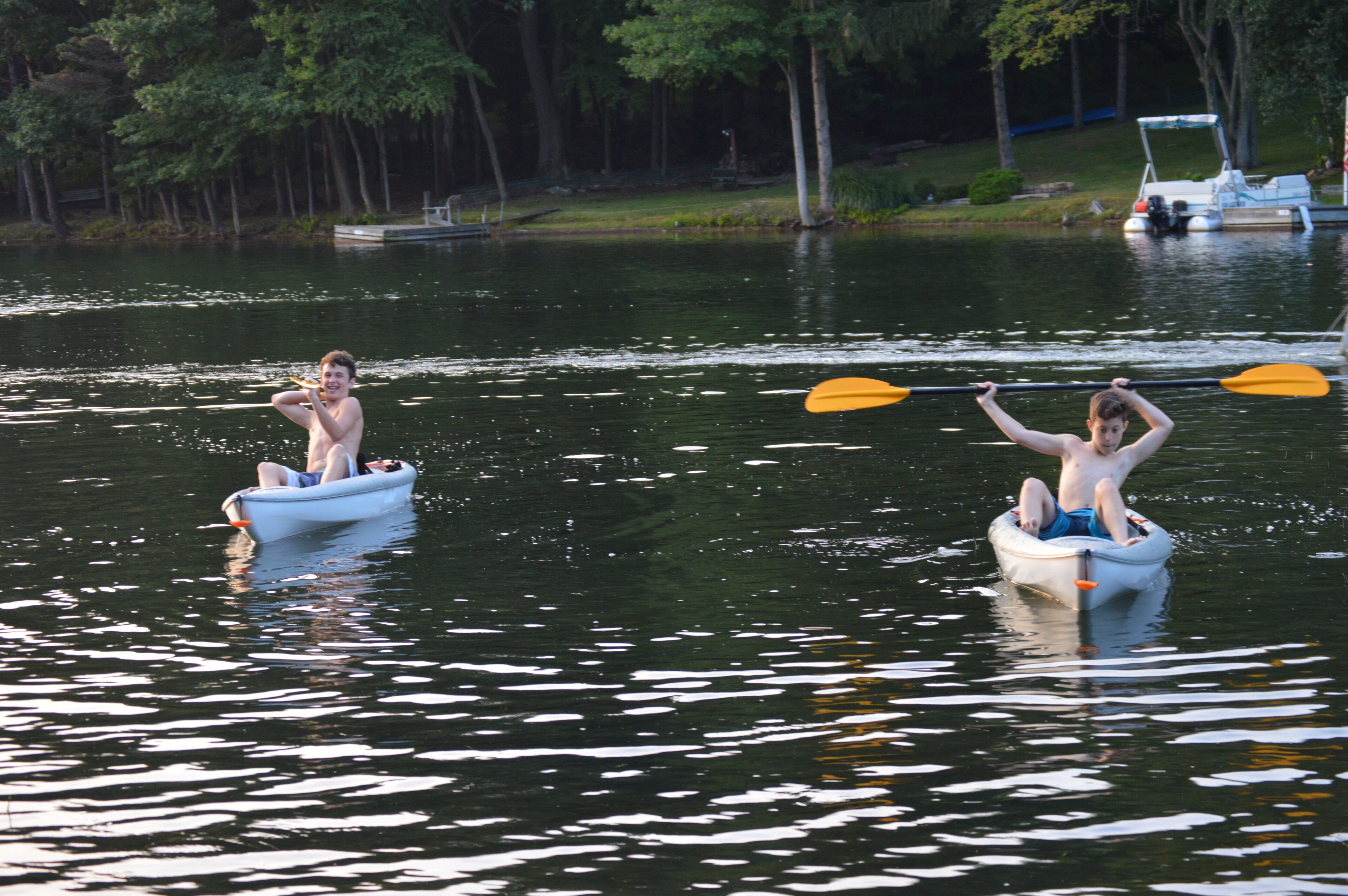 Two people in small boats on a calm lake with trees in the background.