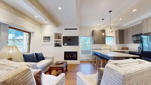 A modern living room with a sofa, coffee table, and striped armchairs. There is a fireplace, kitchen area with bar stools, and a large window. - Christophe Condo 503 - Year-round Outdoor Pool and Hot Tub and Bald Mt. Views (Ketchum)