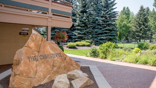 A stone monument with the inscription "THE CHRISTOPHE" in a landscaped garden area with trees and a building in the background. - Christophe Condo 503 - Year-round Outdoor Pool and Hot Tub and Bald Mt. Views (Ketchum)