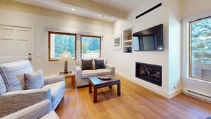 A living room with a sofa, armchair, coffee table, fireplace, TV, and large windows with a view of trees. - Christophe Condo 503 - Year-round Outdoor Pool and Hot Tub and Bald Mt. Views (Ketchum)
