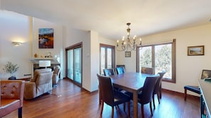 A dining area with a wooden table and chairs, a chandelier, and large windows with a view of trees.