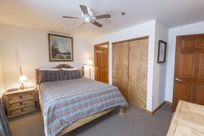 Een slaapkamer met een bed, een houten garderobe, een plafondventilator, een nachtkastje en een lamp.