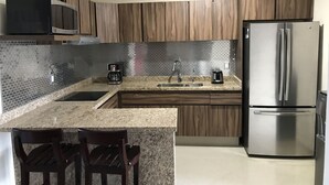 Une cuisine moderne dotée de plans de travail en granit, d’un réfrigérateur en acier inoxydable, d’armoires en bois et d’un dosseret carrelé.