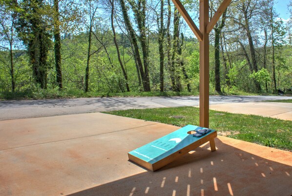 Un banco de madera con cojín azul y una estructura de madera en un parque con sendero de concreto y árboles al fondo.