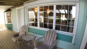 Eine Holzterrasse mit zwei Adirondack-Stühlen und einem kleinen Tisch, ein großes Fenster mit vielen Scheiben und eine Tür mit grüner Umrandung.