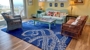 Ein Wohnzimmer mit einem blauen Teppich in Form eines Oktopus, Korbstühlen, einem hellblauen Sofa und einem Holztisch.