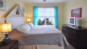 Ein Schlafzimmer mit Bett, Deckenventilator, Fenster mit Vorhängen, Kommode mit Fernseher und Wandbildern.