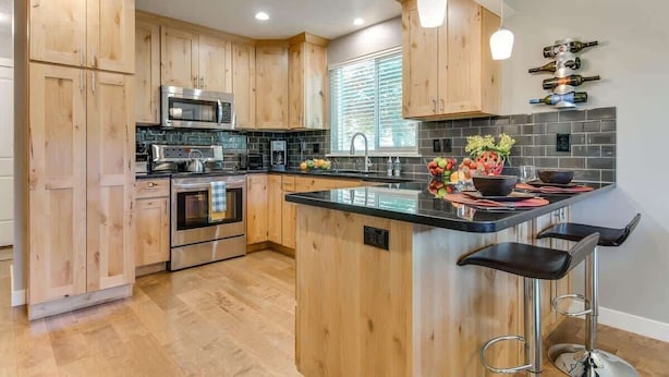 A modern kitchen with wooden cabinets, stainless steel appliances, a central island with bar stools, and a backsplash of black tiles.