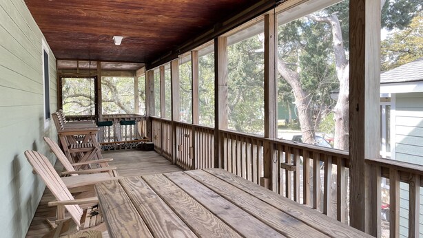 Une véranda couverte avec des bancs en bois et une balançoire, de grandes fenêtres et une vue sur les arbres et une maison.