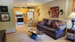 A living room with a brown sofa, a coffee table, and a lamp. There is a kitchen in the background with wooden cabinets and a refrigerator.