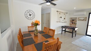 Uma sala de jantar com uma mesa e cadeiras de madeira, um relógio na parede e uma mesa de futebol no fundo.