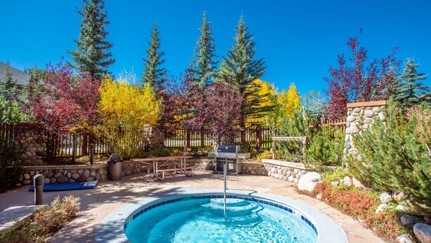 A hot tub surrounded by trees with autumn foliage, a stone wall, and a barbecue grill. - Spacious condo that sleeps 4, steps from the lifts, free wifi & parking (Copper Mountain)