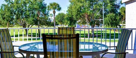A glass-top table with four striped chairs on a balcony with a view of a park and trees.