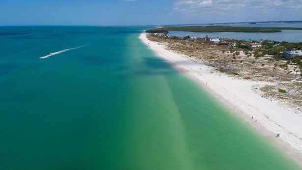 Vista aérea de una playa con aguas turquesas cristalinas, una orilla arenosa y un pequeño poblado con edificios y vegetación.